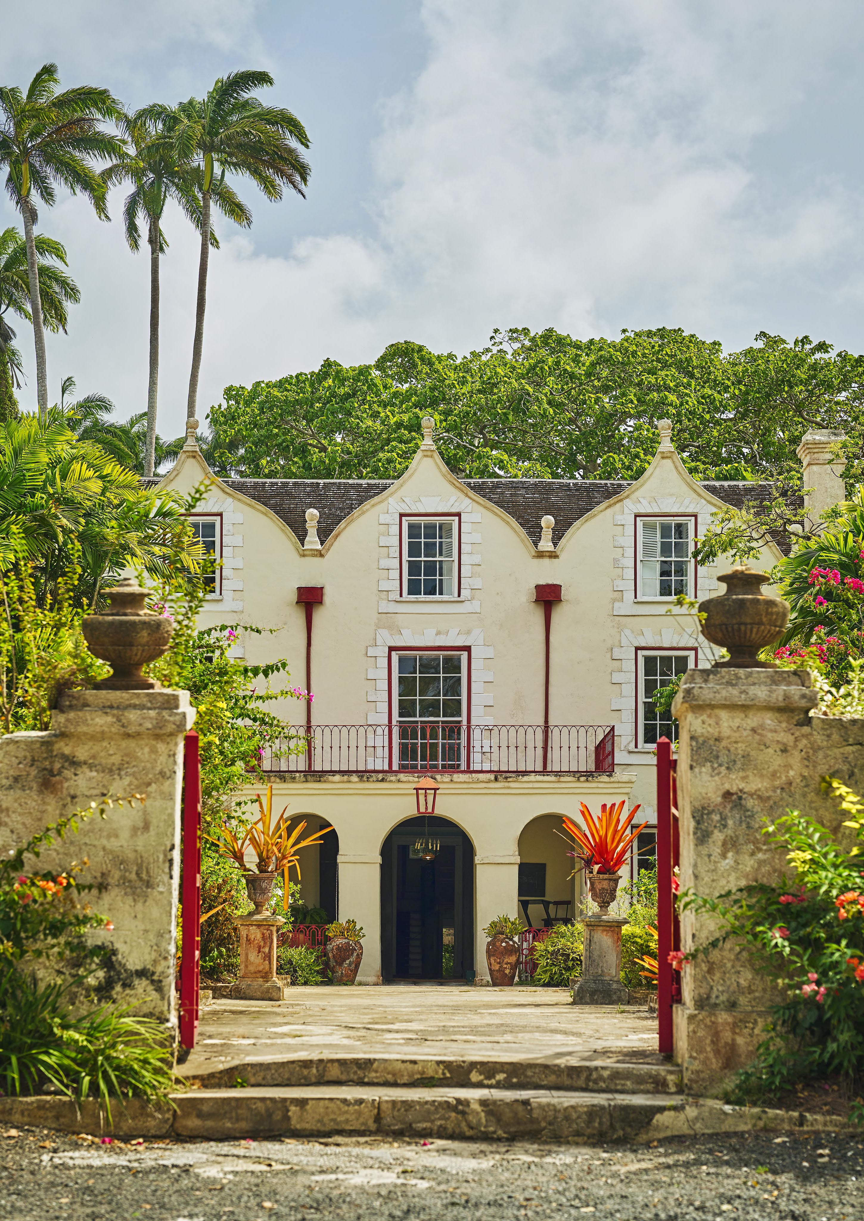 Historic two-story landmark Barbados rum distillery with prominent gables, framed by palm trees and lush greenery. Stone pillars and iron gate mark the entrance.