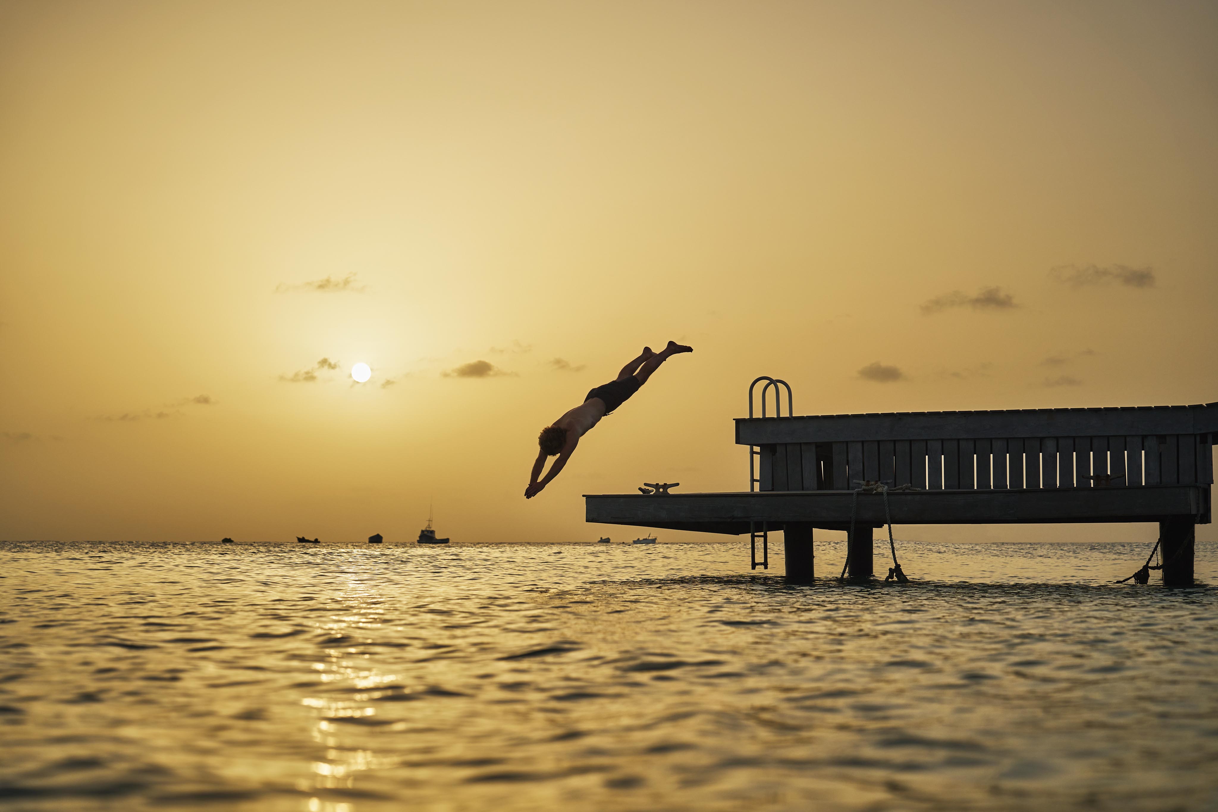 Person diving off a wooden pier into the ocean at sunset, with a golden sky and the sun near the horizon.