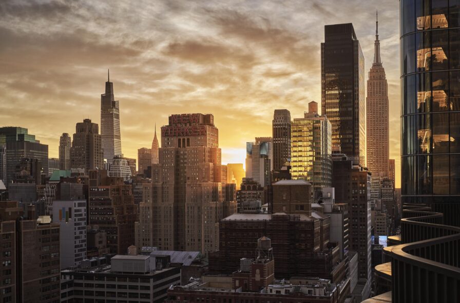 Sunset over a Manhattan city skyline with tall buildings and a cloudy sky. The sun is partially visible, casting a warm glow on the urban landscape.