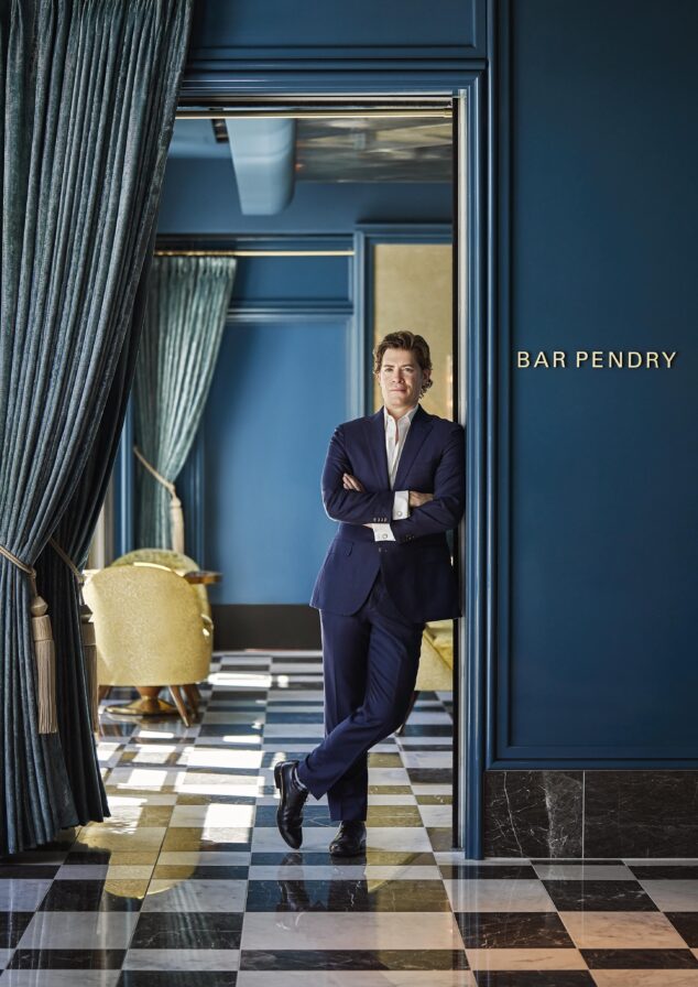 Refined authentic portrait of man in a blue suit leaning against a doorway labeled "Bar Pendry," with elegant blue curtains and checkered floor tiles.