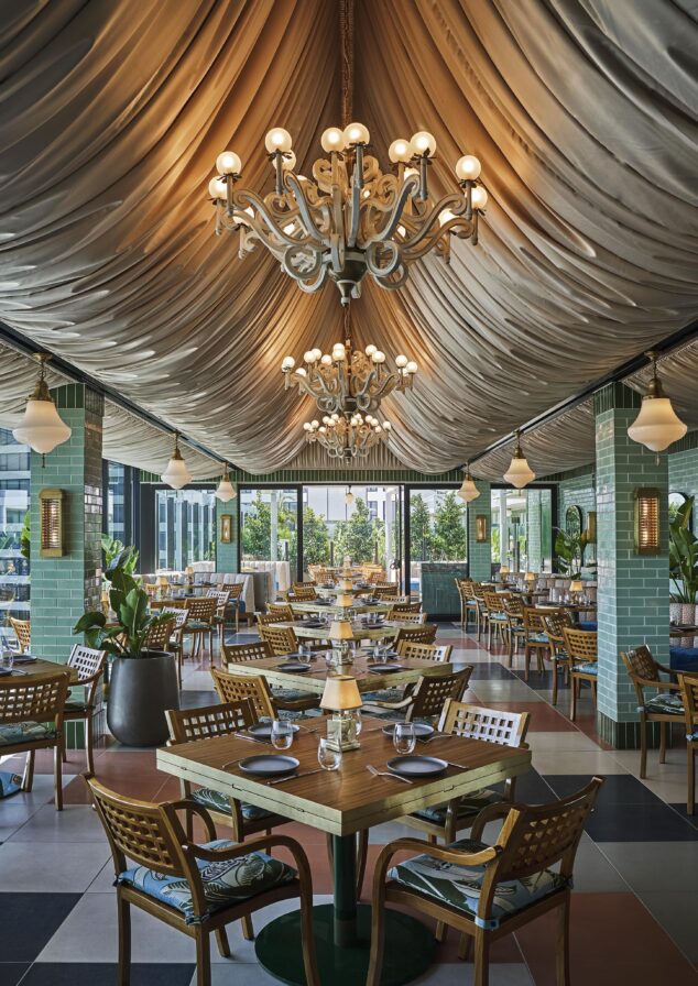 Elegant Pendry West Hollywood restaurant, Merois, interior with draped ceiling, chandeliers, and wooden tables set with plates and glasses. Green tiled columns, checkered floor, and outdoor views in the background.