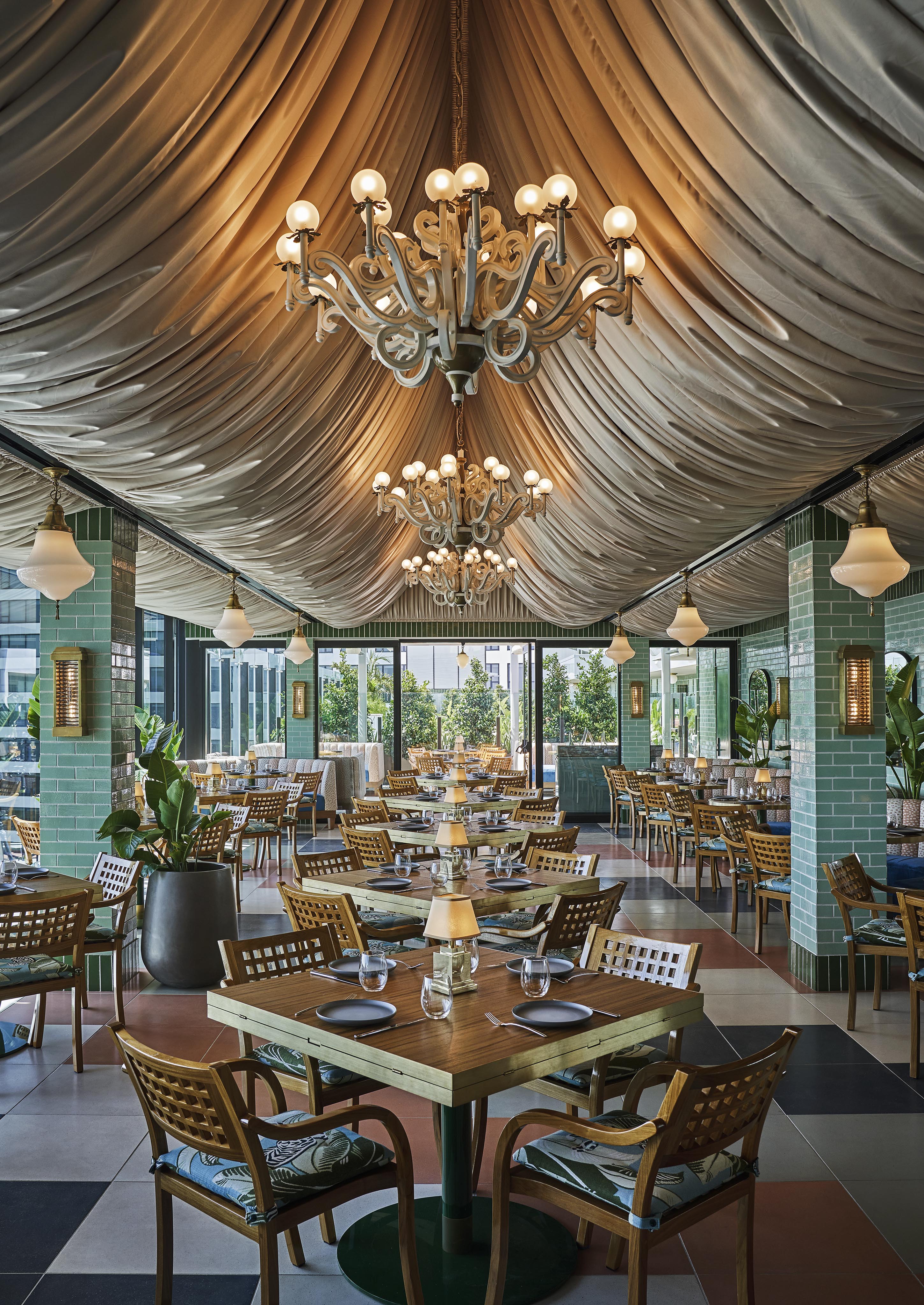 Elegant Pendry West Hollywood restaurant, Merois, interior with draped ceiling, chandeliers, and wooden tables set with plates and glasses. Green tiled columns, checkered floor, and outdoor views in the background.