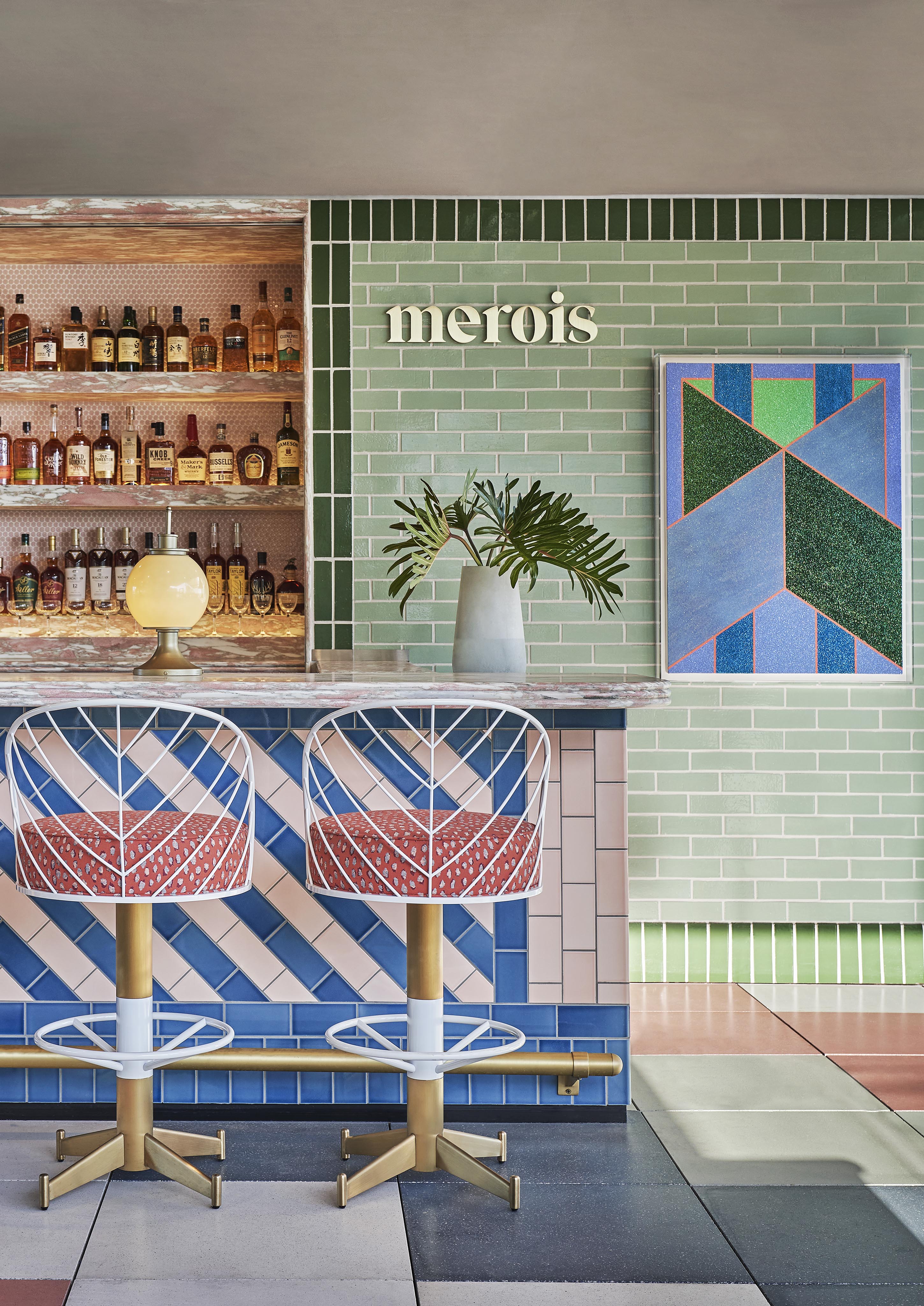 A modern luxury Merois restaurant bar interior within Pendry West Hollywood with geometric tile patterns, two white and red bar stools, a variety of liquor bottles on shelves, and a potted plant on the counter, against a green tiled wall.