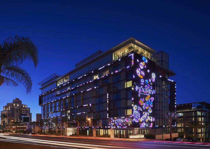 Modern, multi-story Pendry West Hollywood exterior illuminated at night with vibrant, colorful lights and signage reading "Pendry." Cars are visible in motion, creating light streaks on the road.