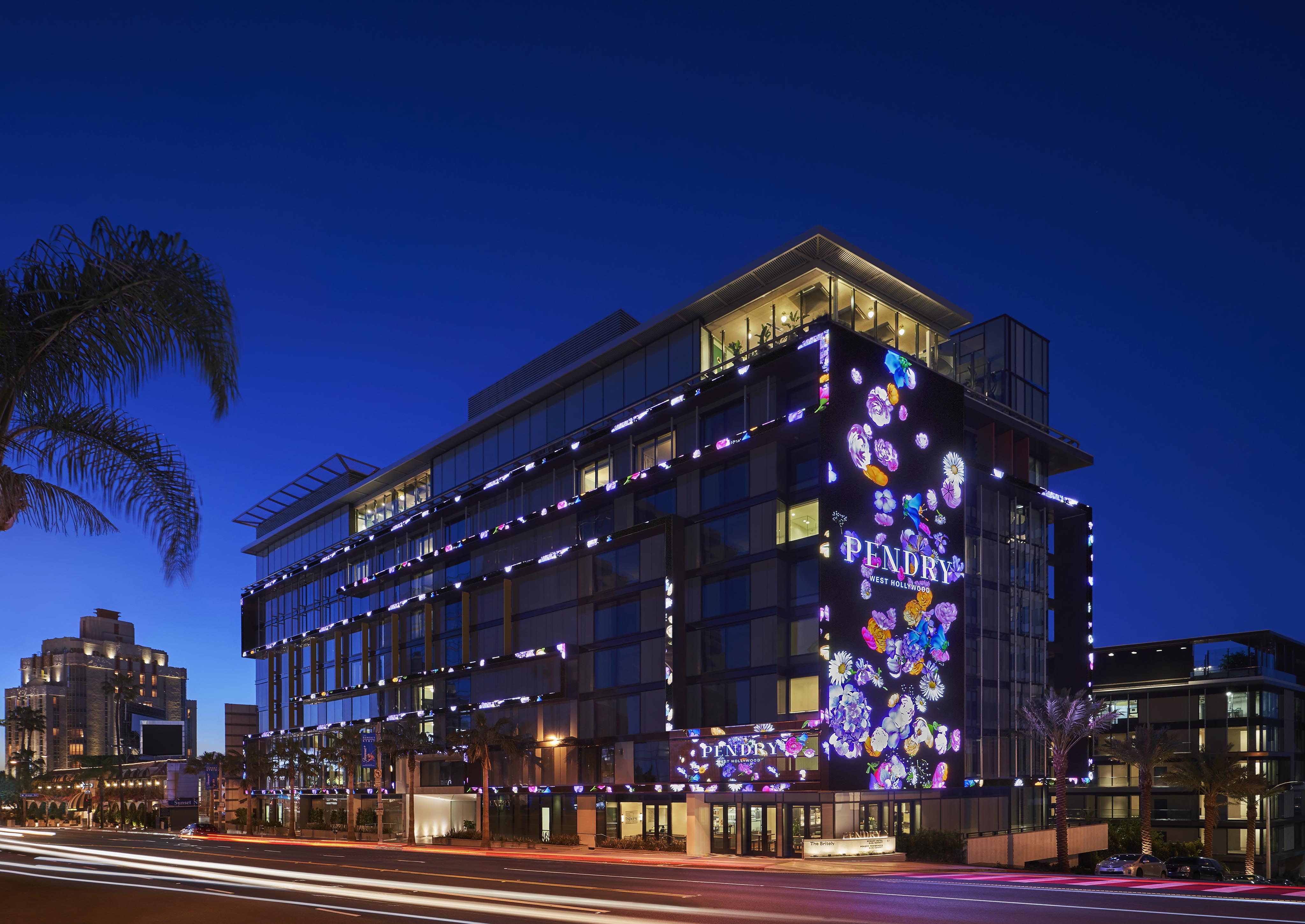 Modern, multi-story Pendry West Hollywood exterior illuminated at night with vibrant, colorful lights and signage reading "Pendry." Cars are visible in motion, creating light streaks on the road.