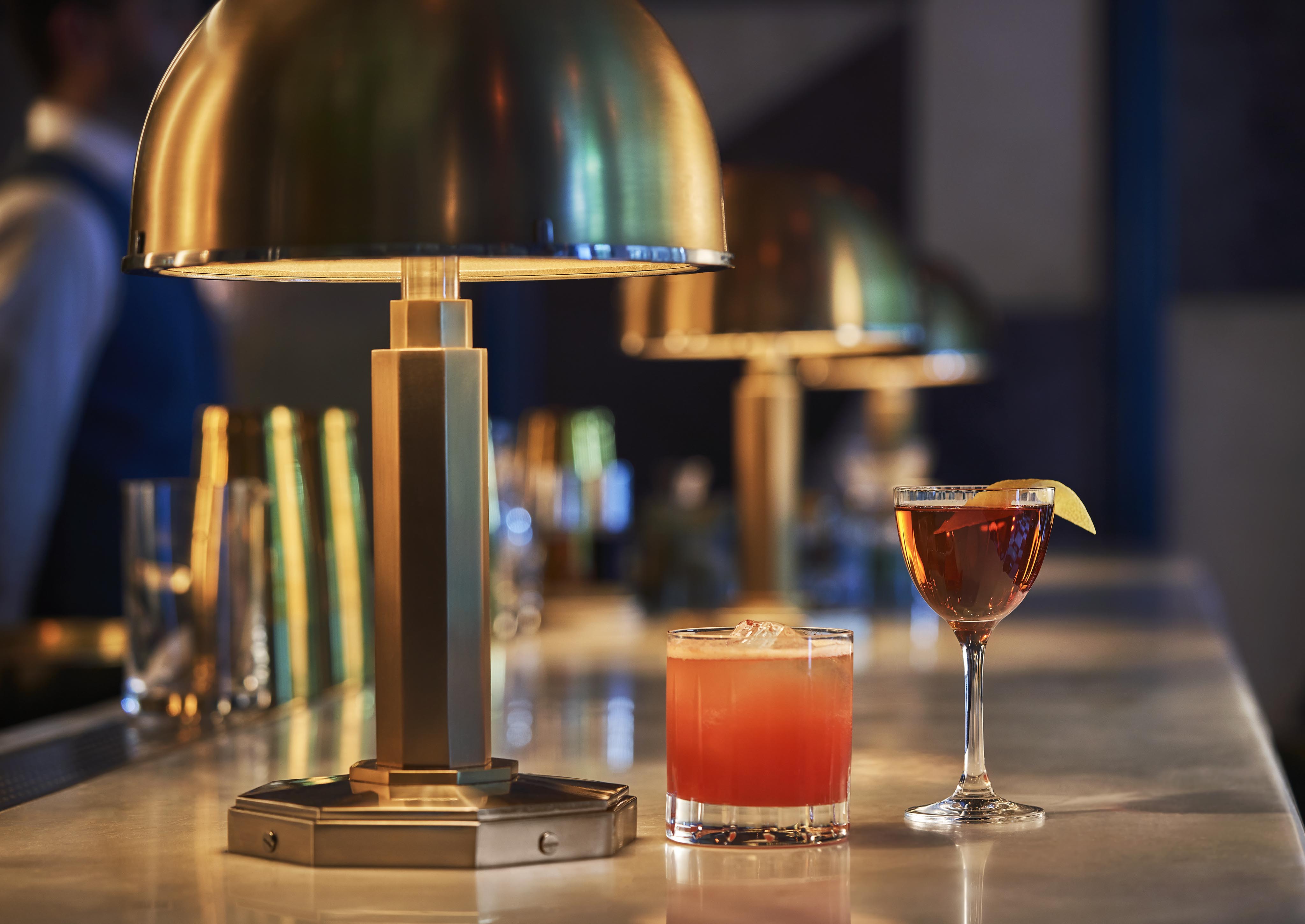 Bar counter with two pristine fine cocktails: a red drink in a short glass with ice, and a dark drink in a stemmed glass with a lemon twist. Brass lamps are in the background. served from the famed Bar Pendry, West Hollywood.