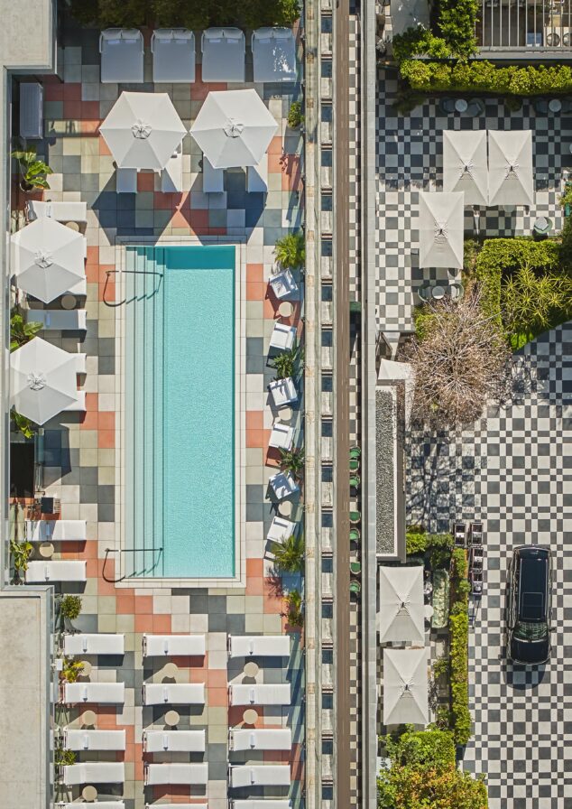 Aerial view of a rectangular pool surrounded by lounge chairs and umbrellas, with a checkered Pendry West Hollywood patio and lobby entrance beneath.