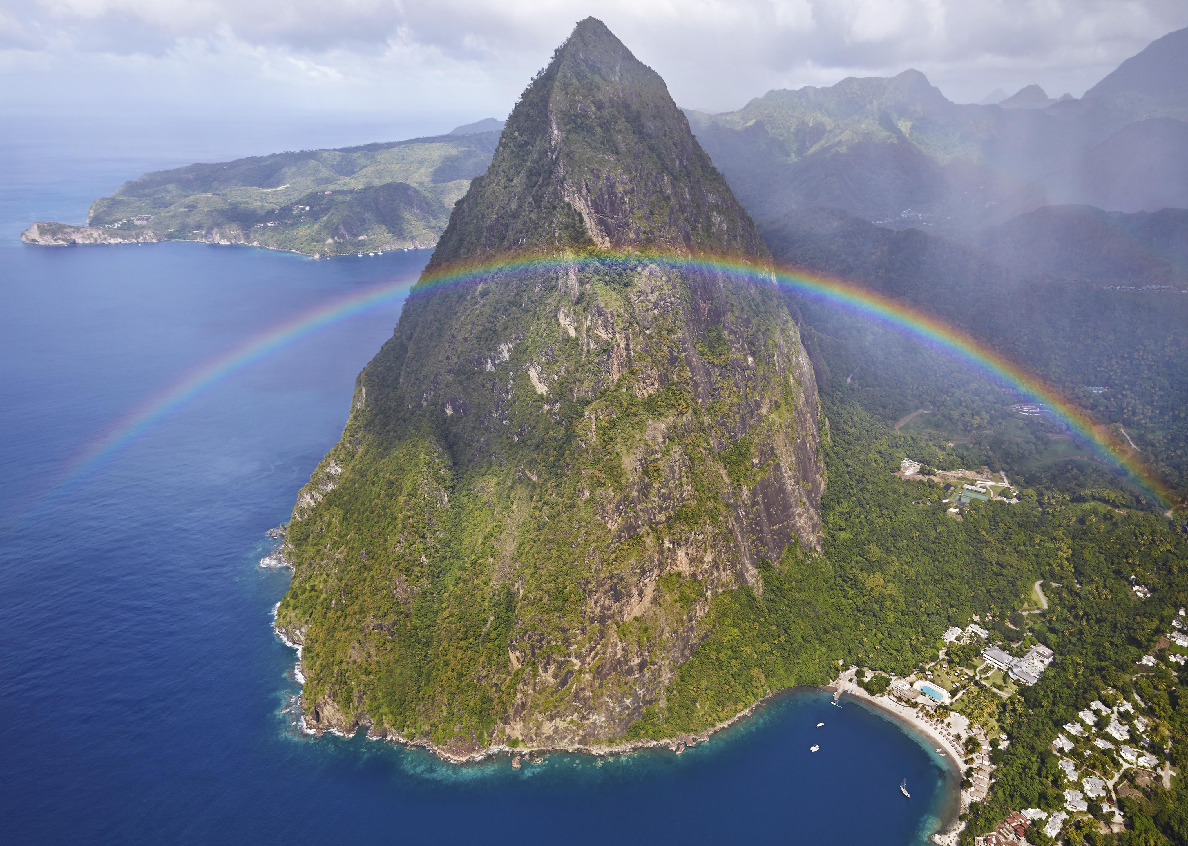 Aerial view of a lush, mountainous landscape with a prominent peak and a vivid rainbow arching over it, meeting the blue ocean below. The scene includes a coastal area with structures.