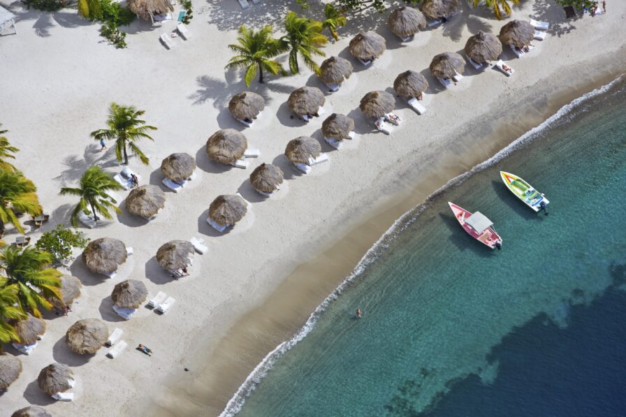 Aerial view of a beach with rows of thatched umbrellas and lounge chairs. Two boats are anchored near the shoreline in the clear water. Palm trees line the sand.