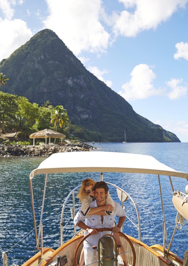 A couple embraces on a boat under a canopy, with lush mountains and a blue sky in the background.