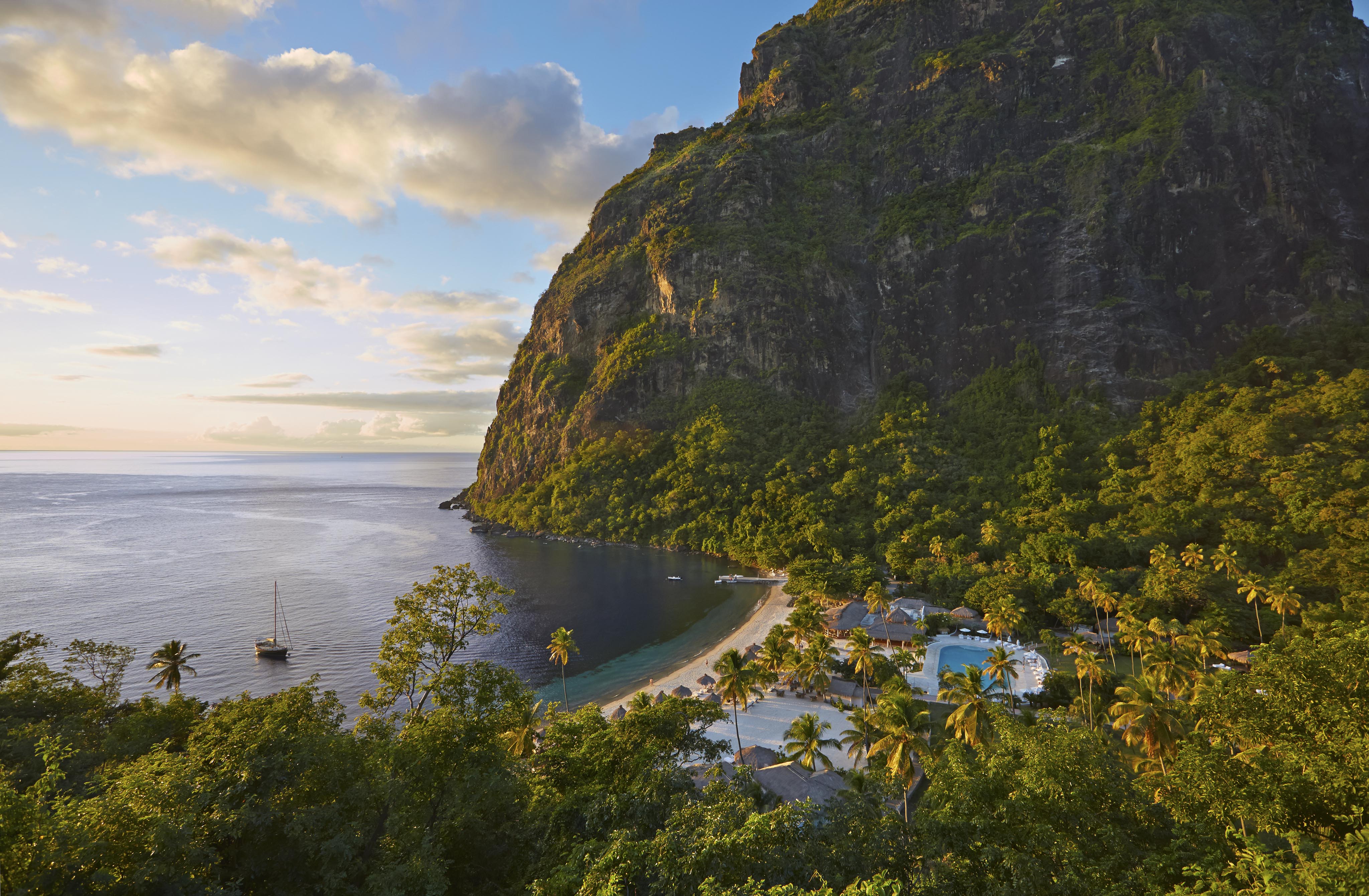 Aerial view of a tropical bay with a sandy beach, lush greenery, a steep mountain, and a sailboat on the water under a cloudy sky.
