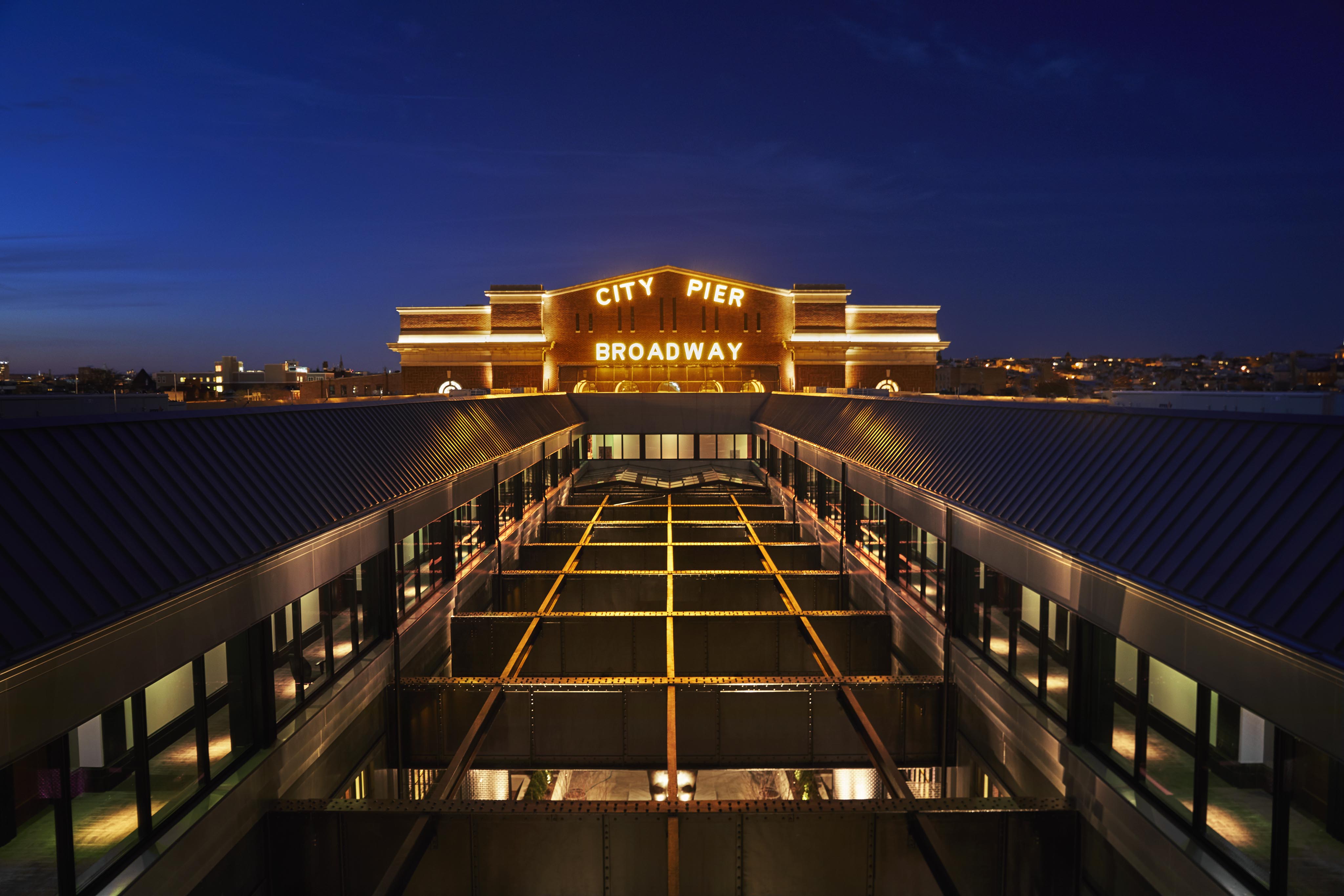 View of a building at night with a lit "City Pier Broadway" sign on the roof, featuring a modern design and rows of windows.