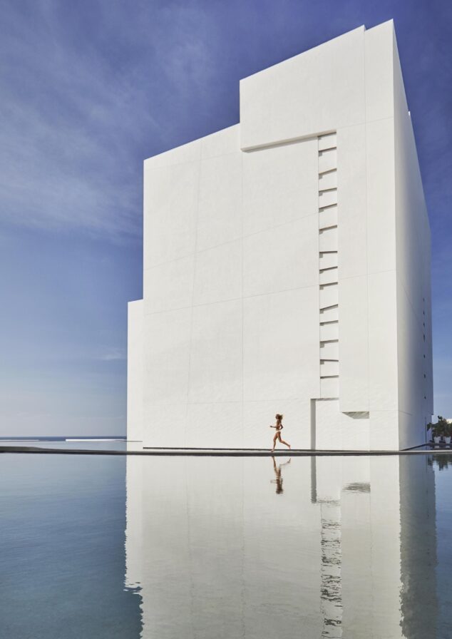 Woman running past large scale white structure surrounded by water, clear Los Cabos blue sky; water reflecting woman and building's mirror image.