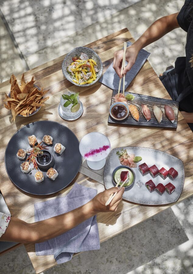 Overhead view of two people dining at a Viceroy Los Cabos restaurant, Nido, table with sushi rolls, nigiri, sashimi, chips, a salad, and two beverages, using chopsticks to pick up food.