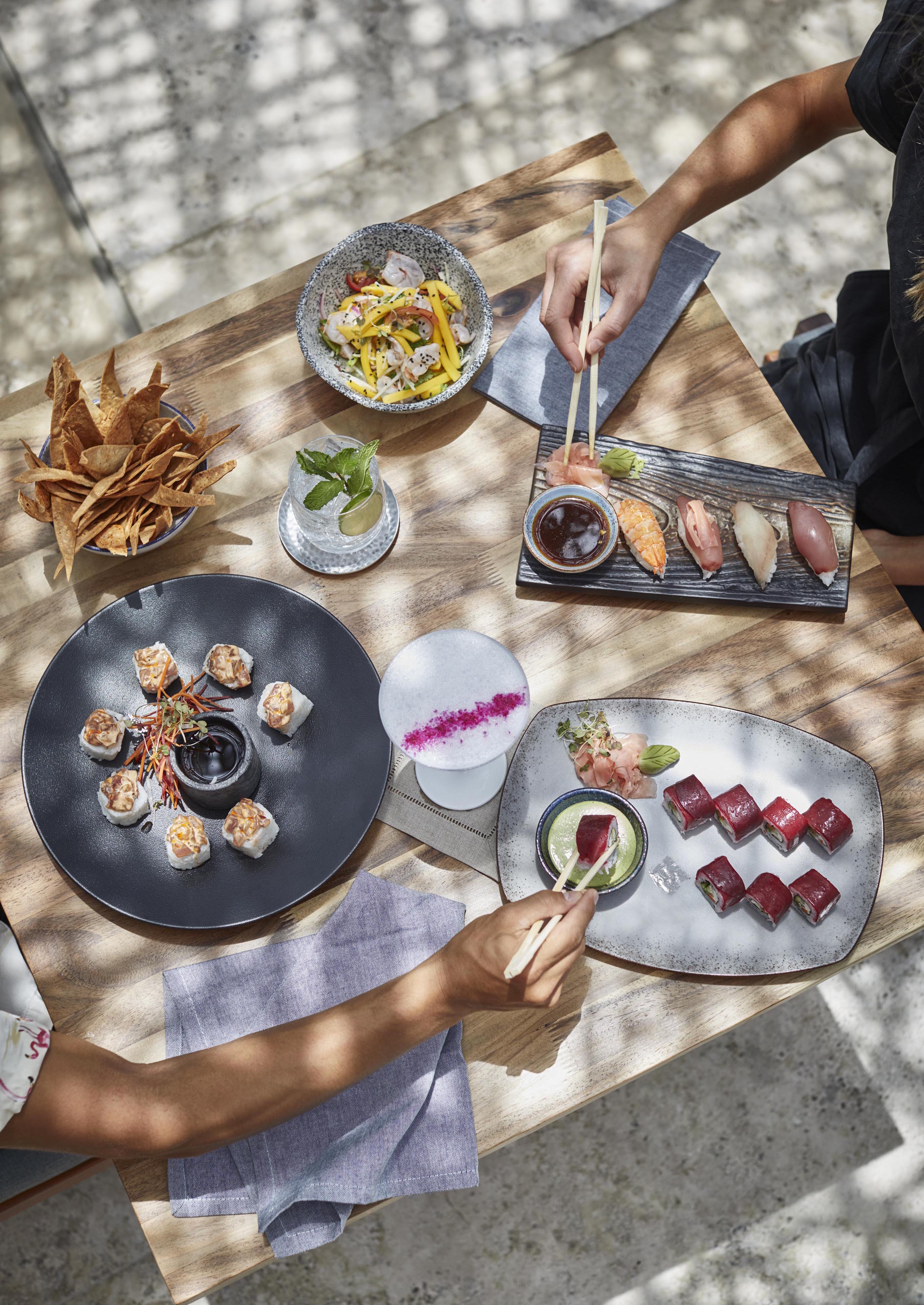 Overhead view of two people dining at a Viceroy Los Cabos restaurant, Nido, table with sushi rolls, nigiri, sashimi, chips, a salad, and two beverages, using chopsticks to pick up food.