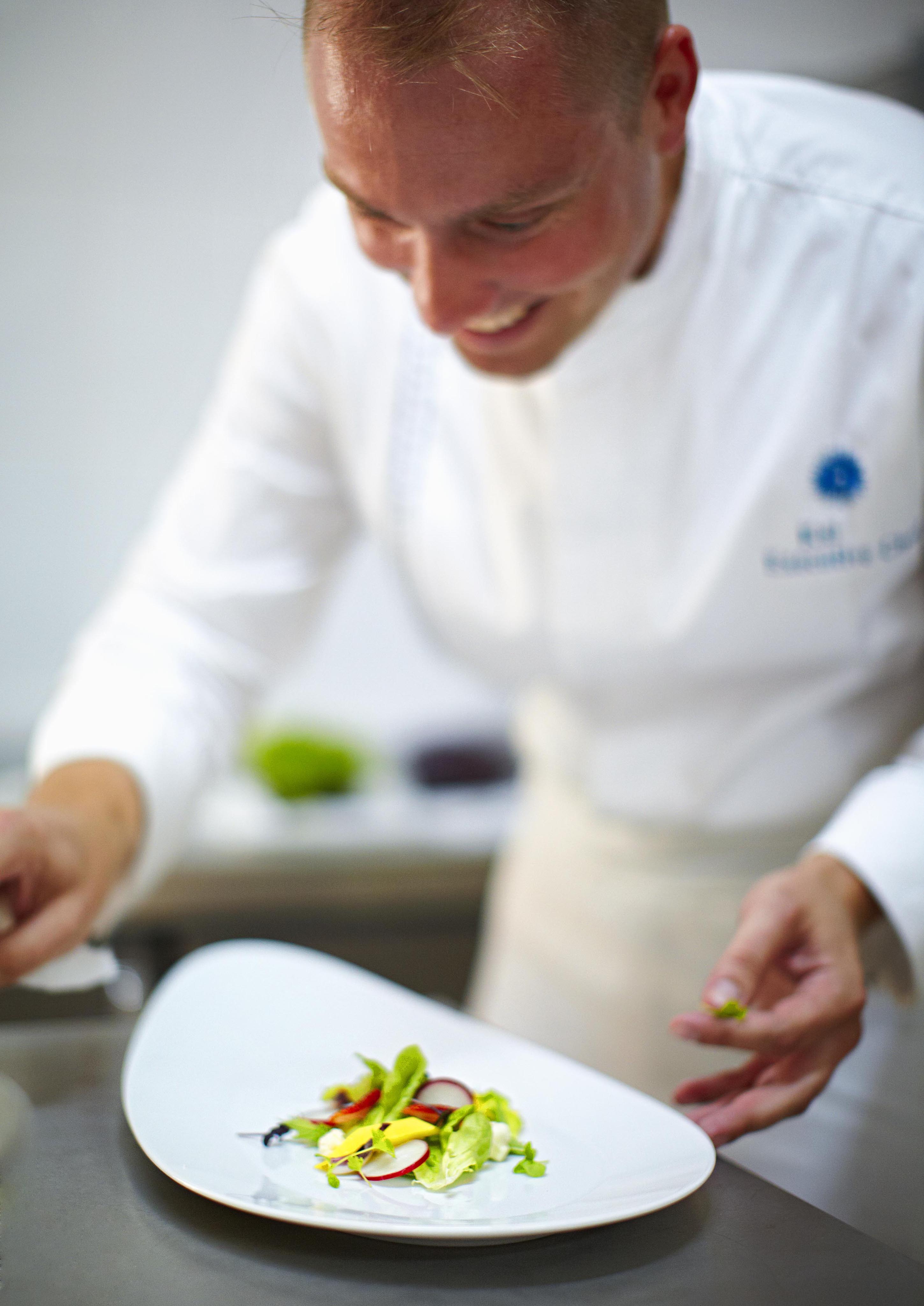 Chef in white uniform plating a dish with vegetables on a white plate, focused and smiling.