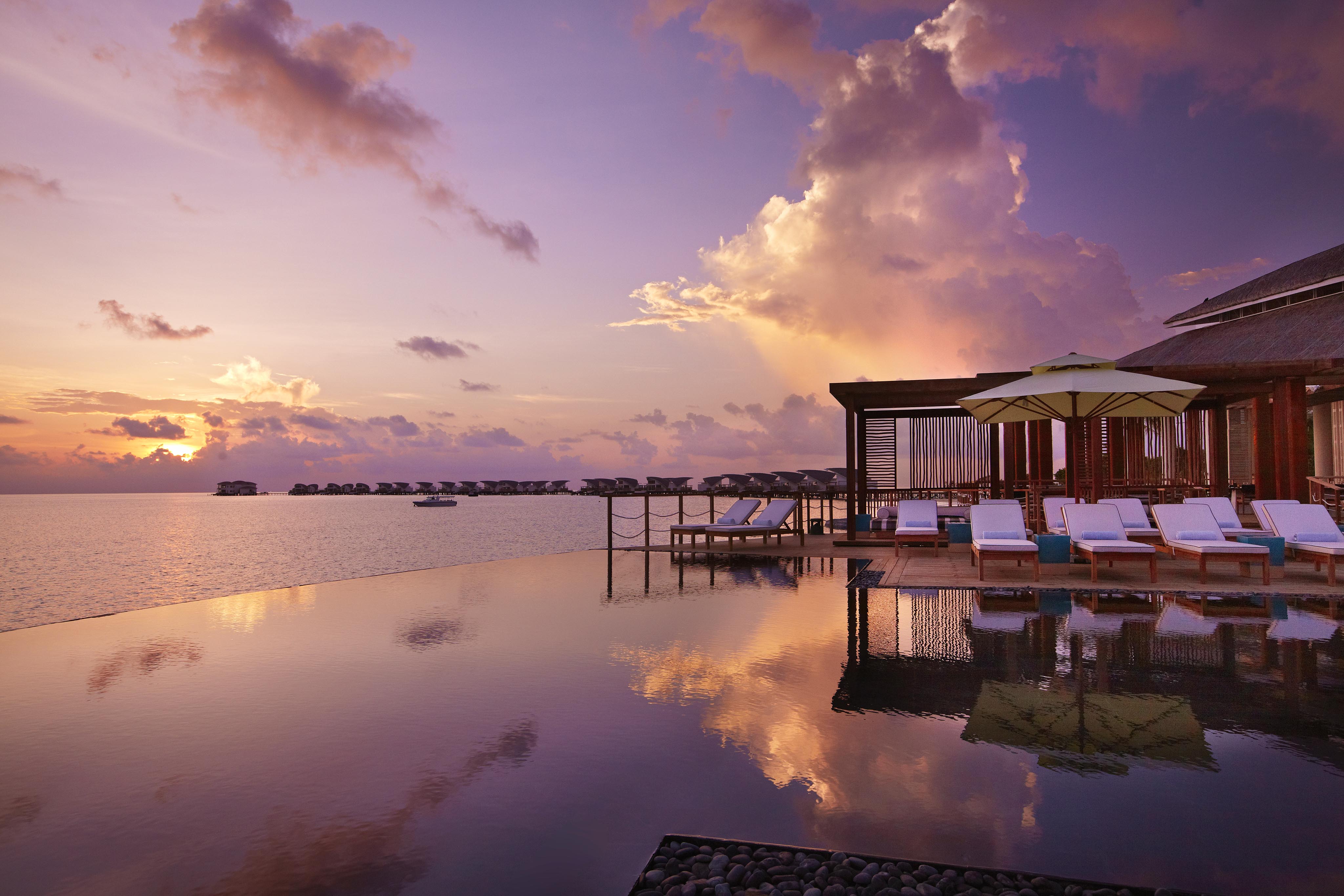 Infinity pool reflecting sunset near beachfront cabanas. Soft orange and purple hues fill the sky, with seating under umbrellas on a deck. Ocean view in the background.