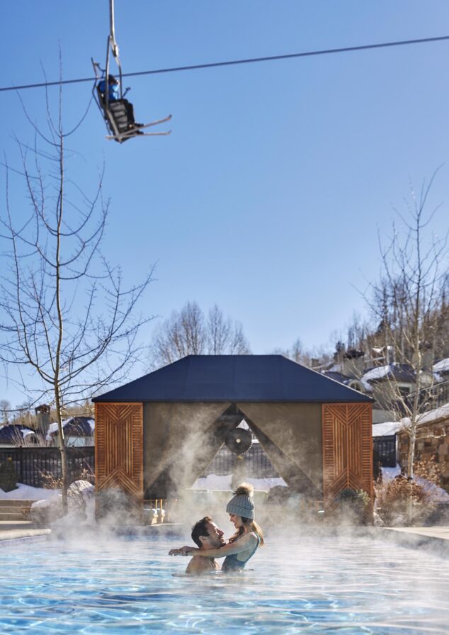 Romantic couple embraced in heated Viceroy Snowmass swimming pool with ski lifts traveling above.