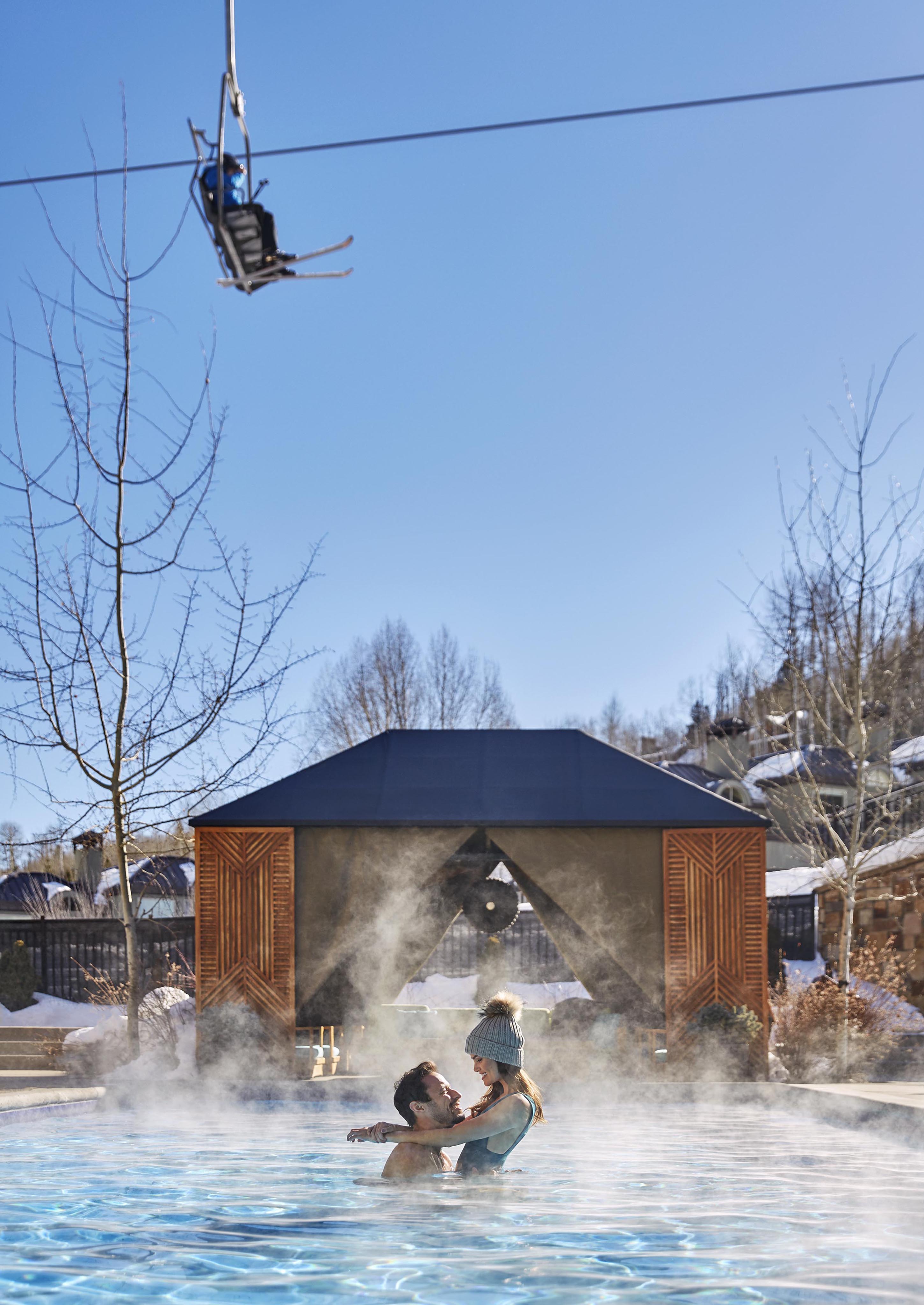 Romantic couple embraced in heated Viceroy Snowmass swimming pool with ski lifts traveling above.