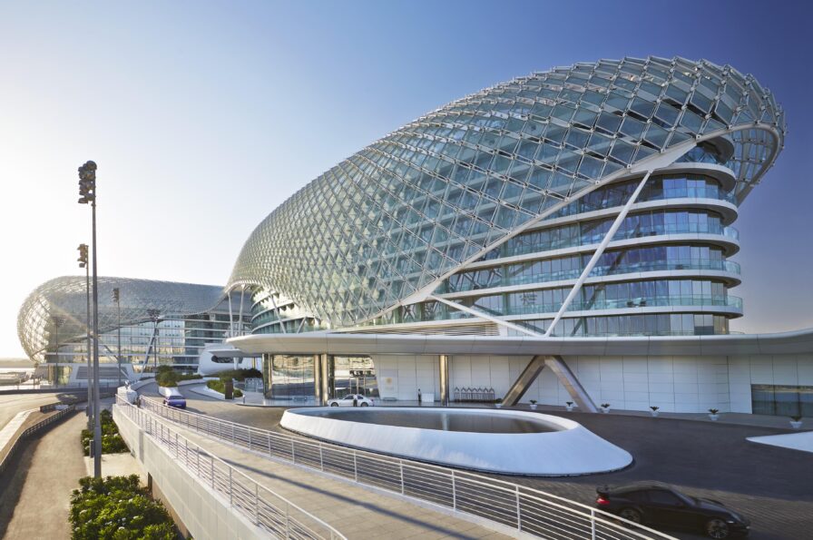 A modern, glass-covered luxury hotel and resort on Yas Island UAE with curved architecture and a mesh-like exterior, captured in stunning Middle East photography, stands beside a road under a clear sky.