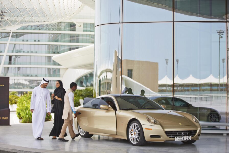 Three people stand near a gold Ferrari parked outside a modern glass building, its windows reflecting striking architecture—perfect for luxury hotel and resort photography in the Middle East.