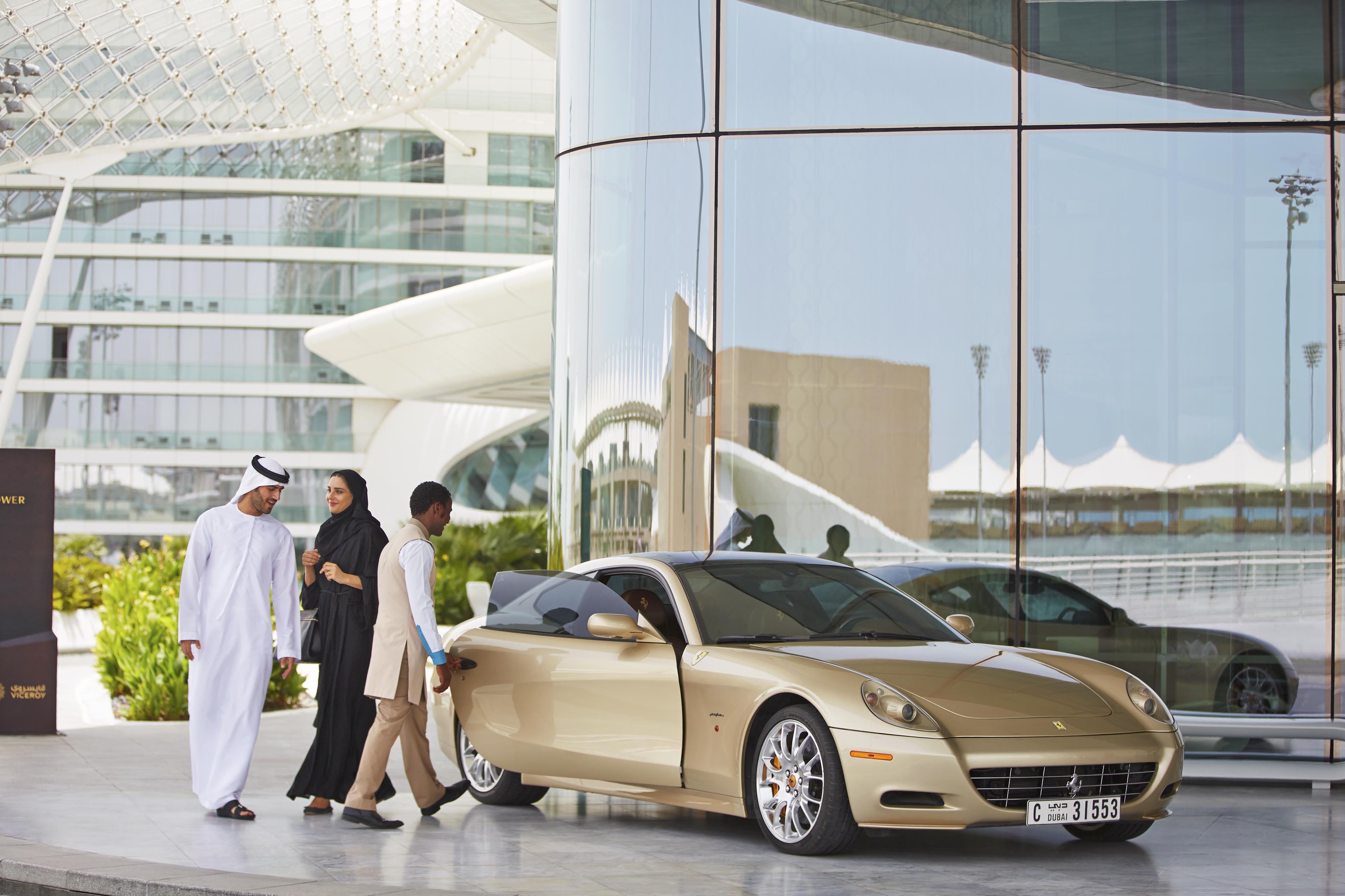 Three people stand near a gold Ferrari parked outside a modern glass building, its windows reflecting striking architecture—perfect for luxury hotel and resort photography in the Middle East.