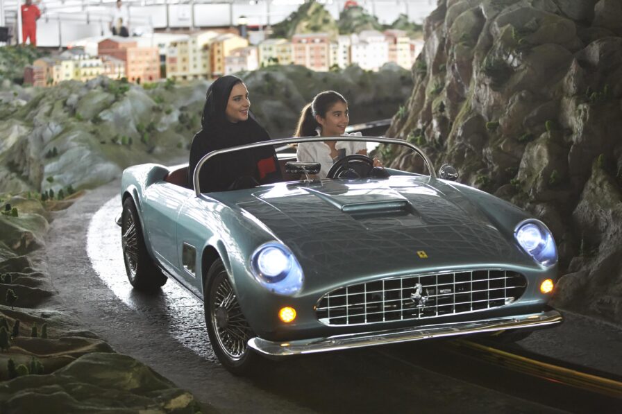 Two women sit in a small blue convertible on a track, surrounded by a model landscape with miniature buildings and mountains within Yas Island Ferrari World, evoking the elegance of luxury hotel and resort photography in the Middle East.