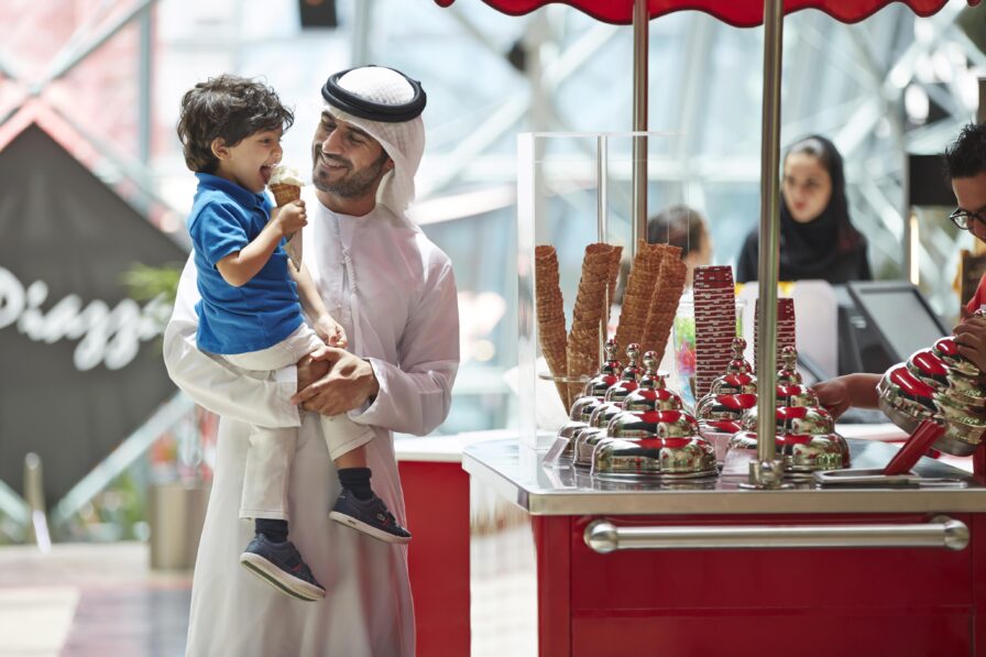 A man in traditional Middle Eastern clothing holds a young boy enjoying an ice cream cone at an ice cream stand, with cones and toppings displayed on the counter—a delightful scene of luxury hotel and resort photography in the Middle East.