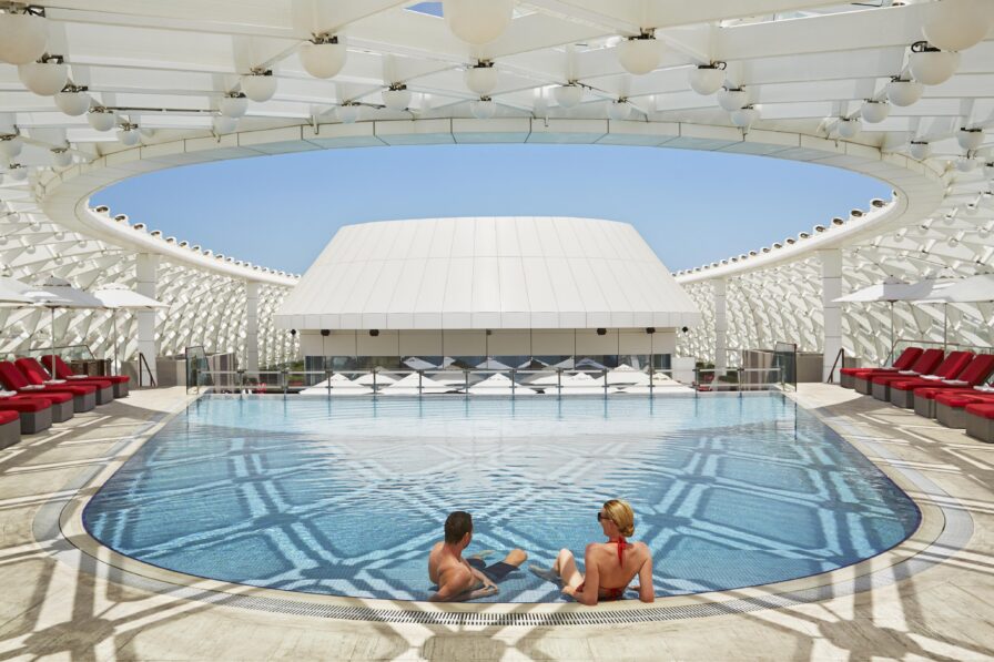 Two people sit at the edge of a circular outdoor Yas Island destination pool, surrounded by red lounge chairs under a modern white canopy—capturing the elegance of luxury hotel and resort photography in the Middle East on a sunny day.