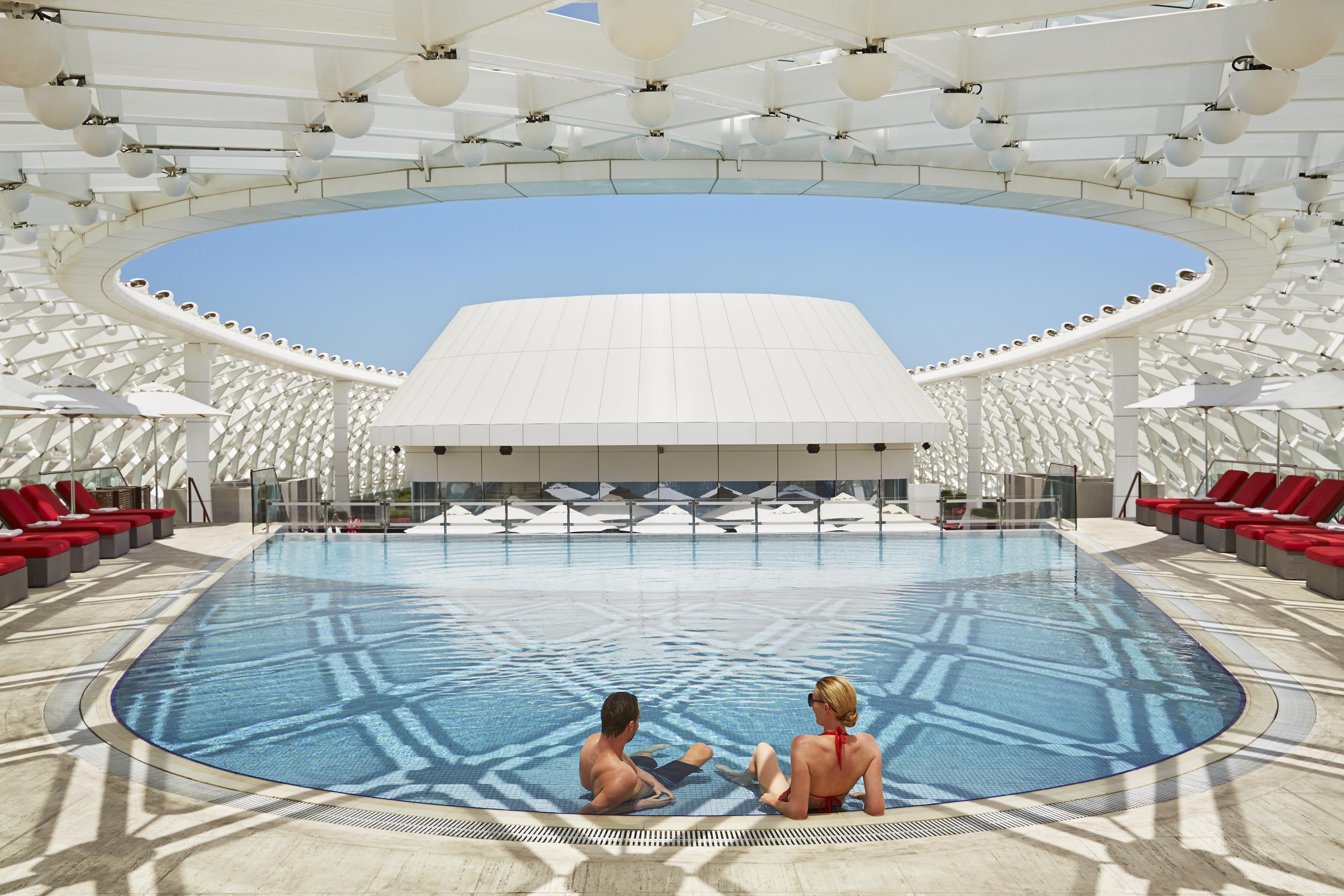 Two people sit at the edge of a circular outdoor Yas Island destination pool, surrounded by red lounge chairs under a modern white canopy—capturing the elegance of luxury hotel and resort photography in the Middle East on a sunny day.