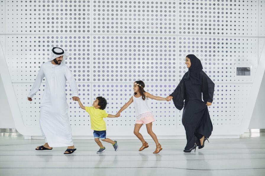 A family of four, two adults and two children, hold hands and walk together indoors in front of a modern, white wall with circular patterns, showcasing luxury hotel and resort photography in the Middle East.
