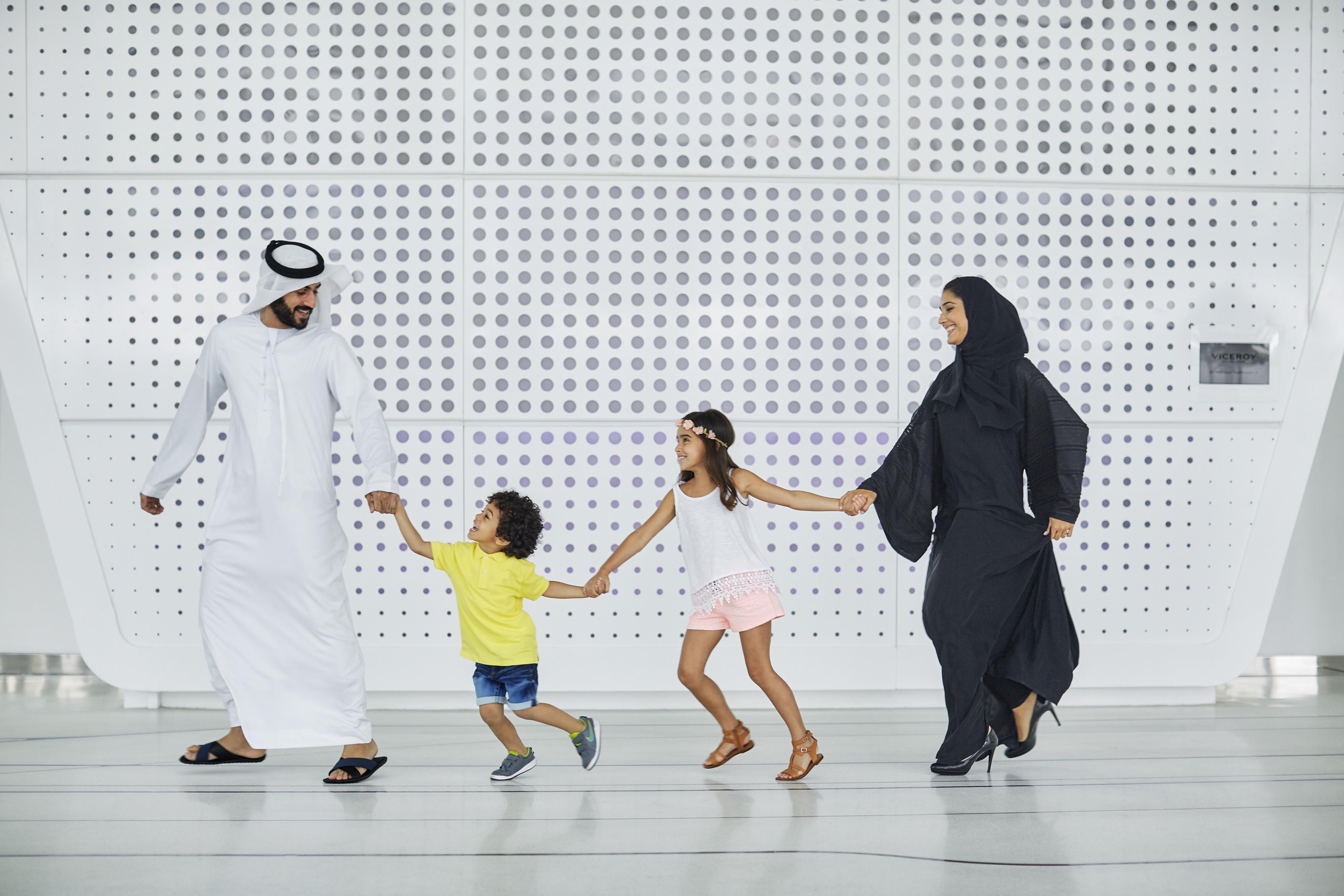 A family of four, two adults and two children, hold hands and walk together indoors in front of a modern, white wall with circular patterns, showcasing luxury hotel and resort photography in the Middle East.