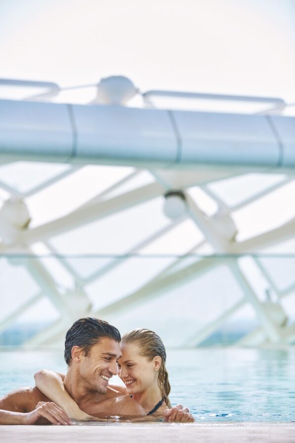 A couple authentically smiles and embraces while leaning on the edge of a pool against a background of modern architecture structure of Viceroy Yas Island luxury hotel.