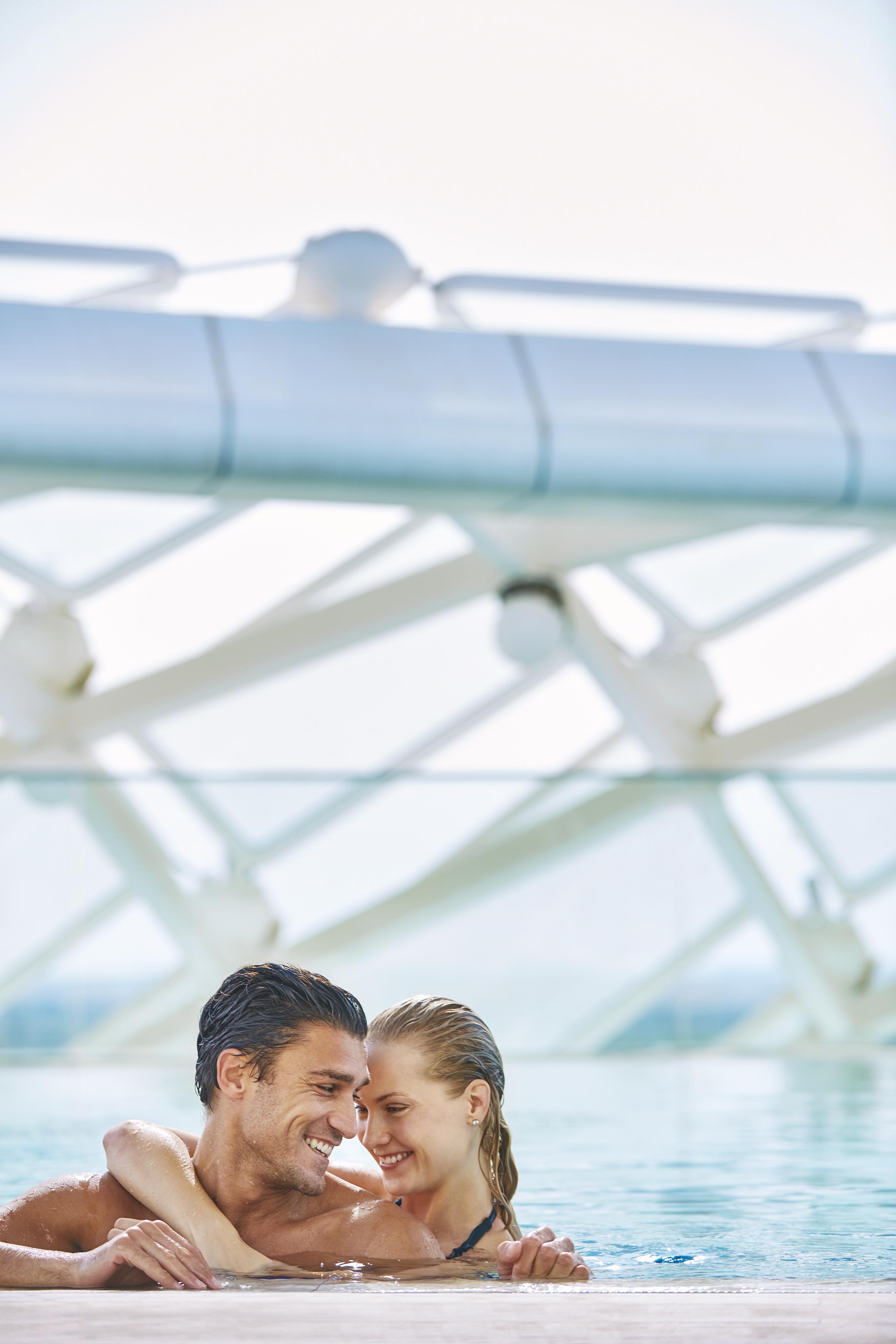 A couple authentically smiles and embraces while leaning on the edge of a pool against a background of modern architecture structure of Viceroy Yas Island luxury hotel.