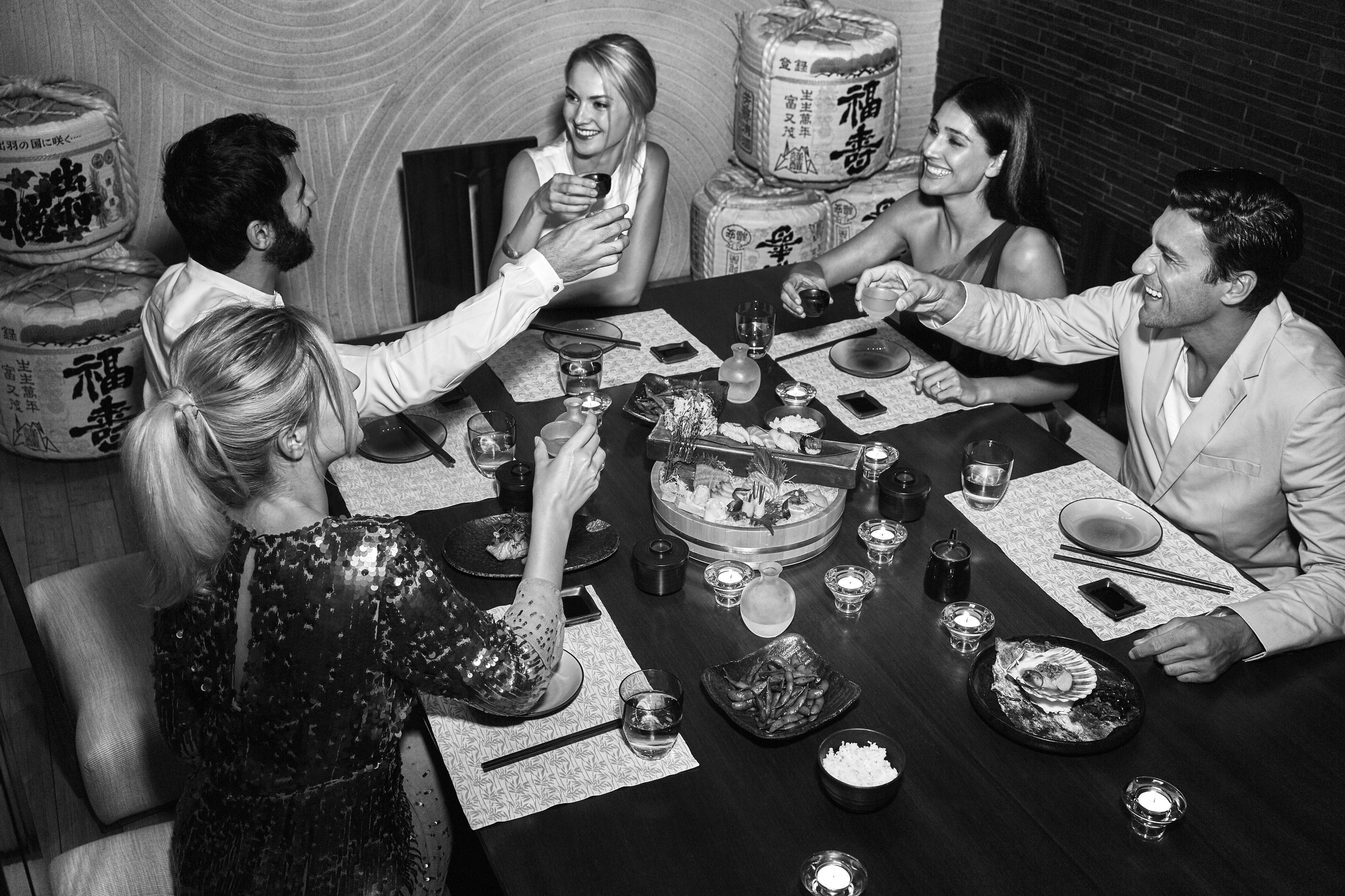 Five people sit around a table in a luxury hotel and resort restaurant in the Middle East, raising glasses in a toast, with sushi and other exquisite dishes before them.