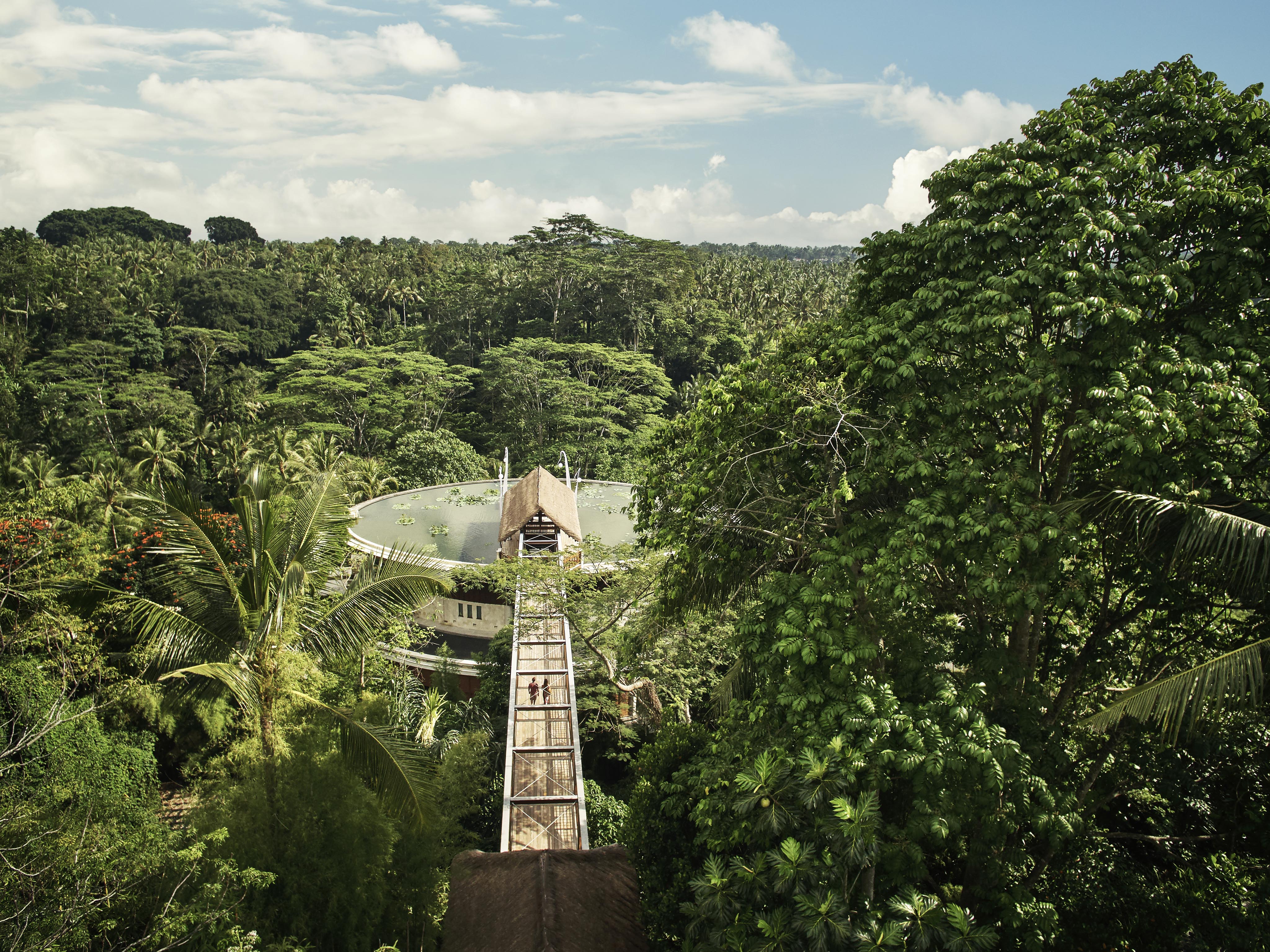 Captured by Christian Horan Photography, an aerial view reveals a long bridge leading to a unique architecture of Four Seasons Bali at Sayan circular building, nestled within a dense green jungle and trees under a partly cloudy sky.