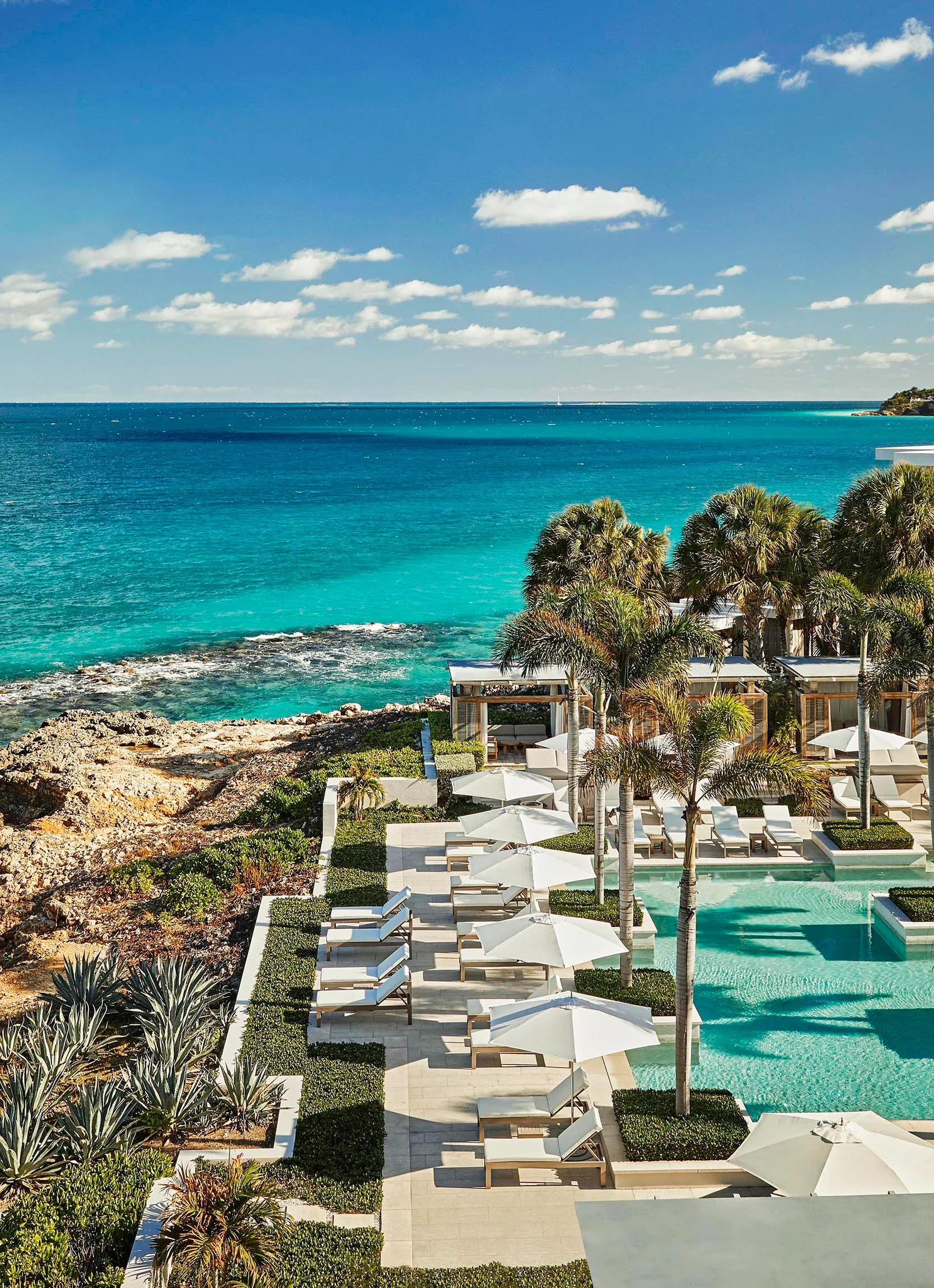 Four Seasons Anguilla resort pool area with umbrellas and lounge chairs, overlooking a rocky coastline and turquoise ocean.