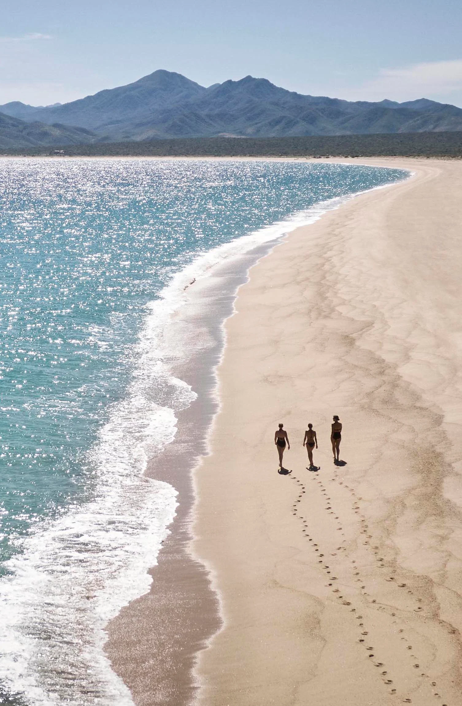 Three people walk along a private Los Cabos beach with gentle waves and mountainous terrain in the background.