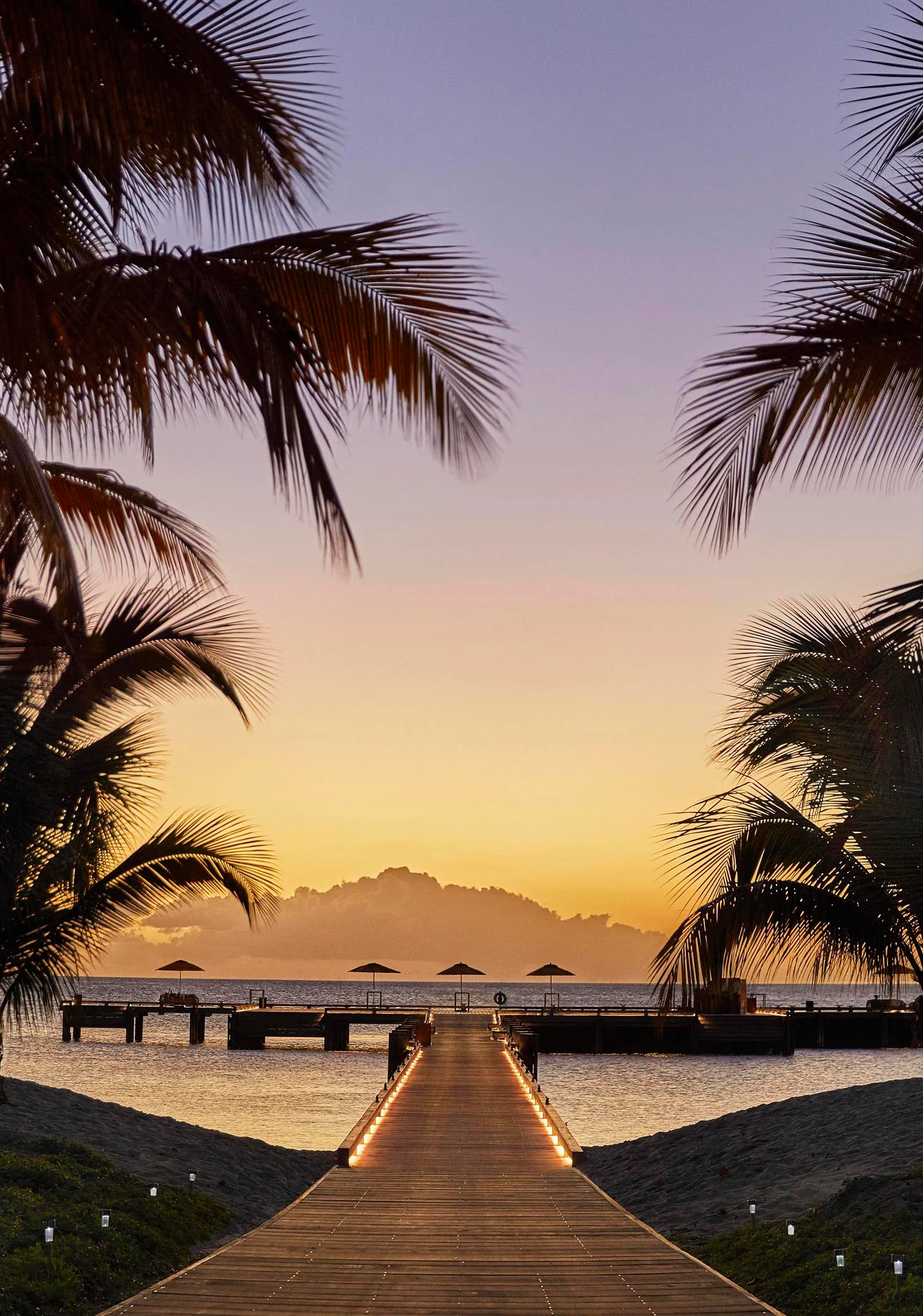 A wooden pier extends over calm Nevis water at sunset, framed by palm trees. Umbrellas are visible at the pier's end, with a distant mountain silhouetted against the sky.
