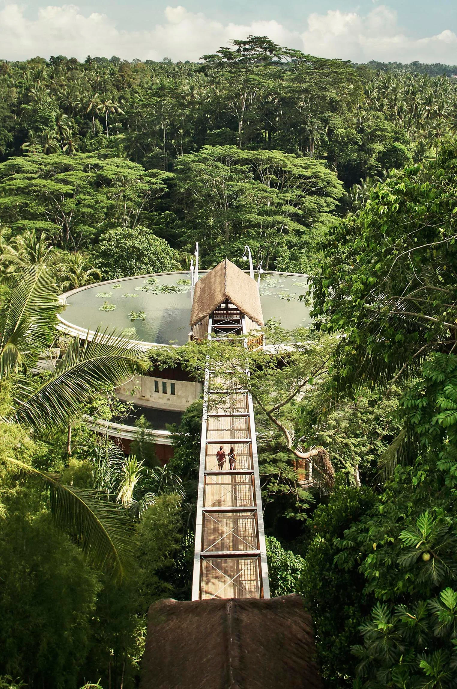 Aerial view of Four Seasons Bali Sayan with long wooden walkway leading to a thatched-roof structure surrounded by dense tropical forest, with a circular building partially visible below—a scene beautifully captured by Christian Horan Photography.