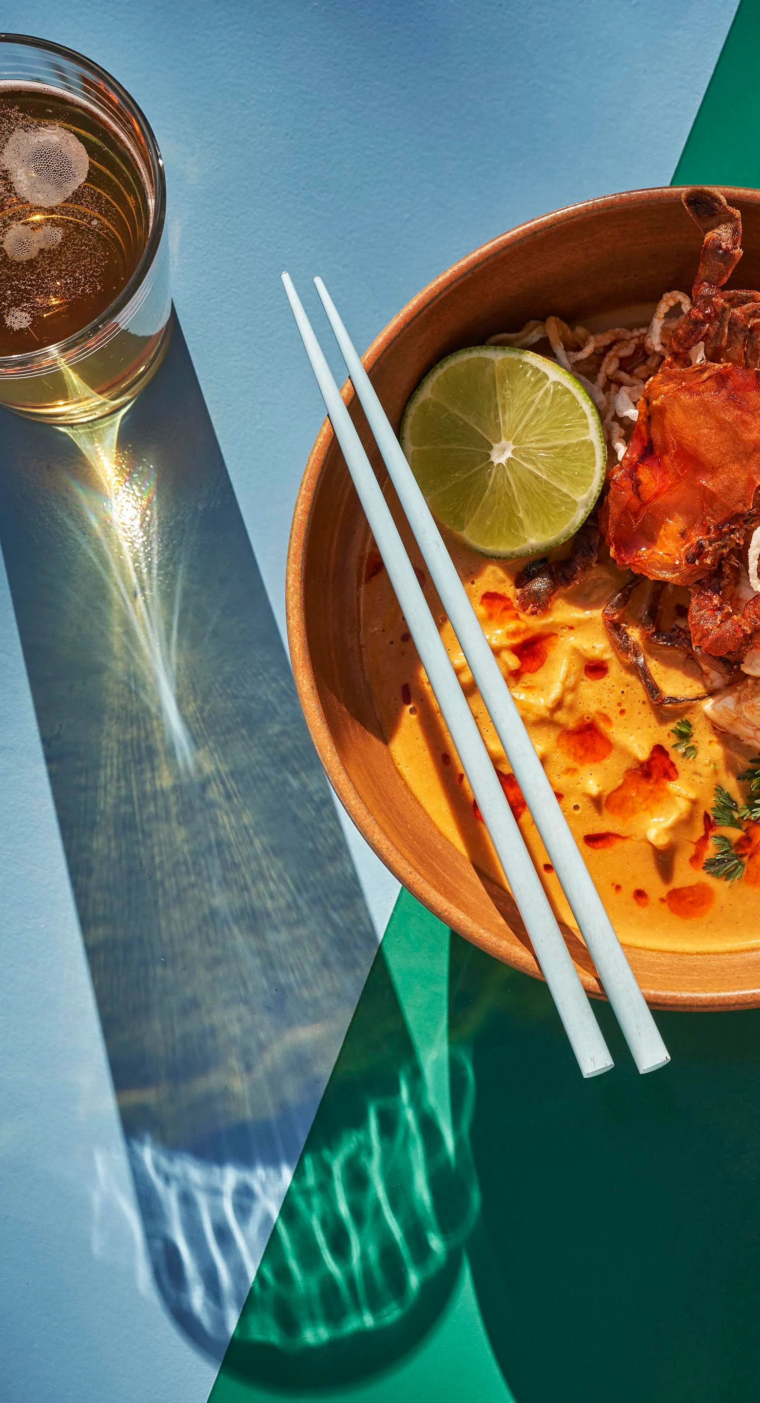 Elevated top down view of bowl of spicy ramen with sliced lime and crispy topping, paired with a glass of iced tea. Chopsticks rest on the bowl. Shadows cast on a blue and green surface.